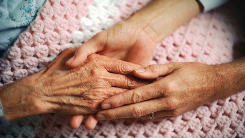 A person holds the hand of an elderly woman, who is covered with a pink and white crocheted blanket.