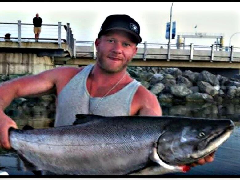 A muscular young white man in a grey tank top and black baseball cap stands in front of a pier holding a very large salmon.