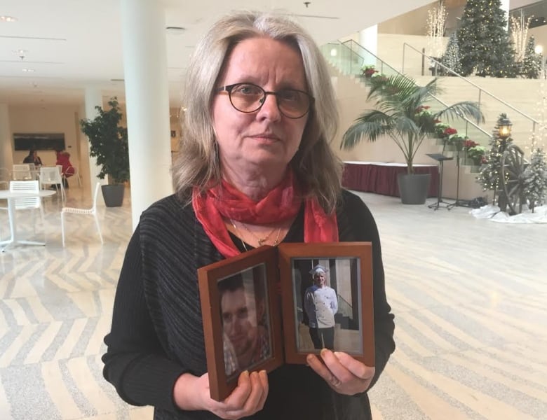 An older woman wearing glasses has a neautral expression while holding a picture frame of her son.