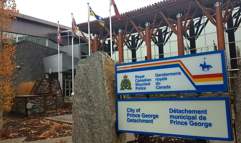 A sign announcing the City of Prince George RCMP detachment is seen next to a stone pillar outside a glass-walled building with trees showing their fall colours.