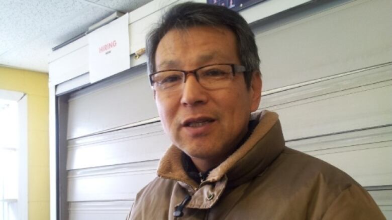 A man with dark hair and square glasses smiles at the camera. He is dressed in a light brown coat and appears to be behind the cash of a convenience store. 