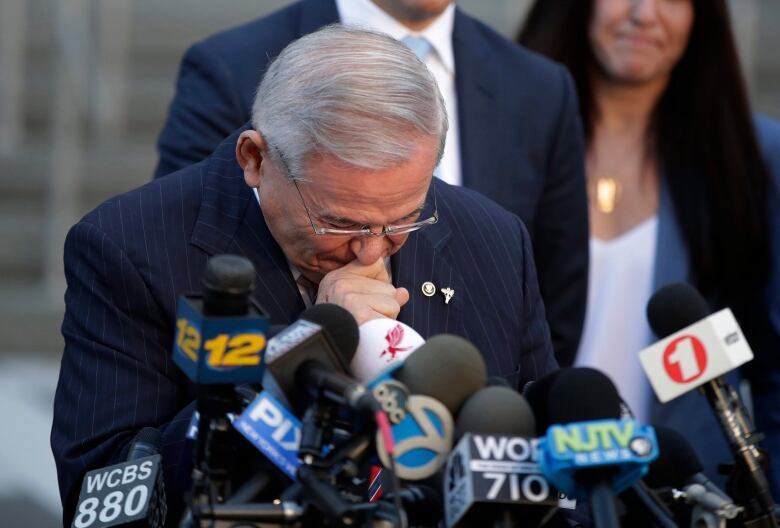 A man grasps his hands, head bowed, in front of several microphones.