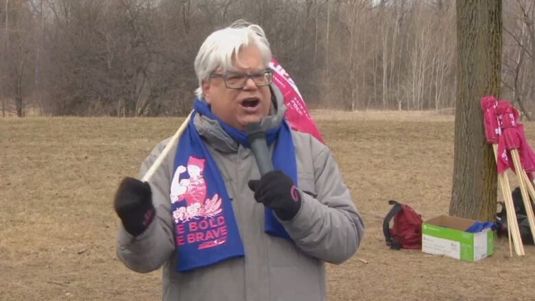 A man with white hair and glasses wearing a grey coat and a scarf holds a flag and a microphone.