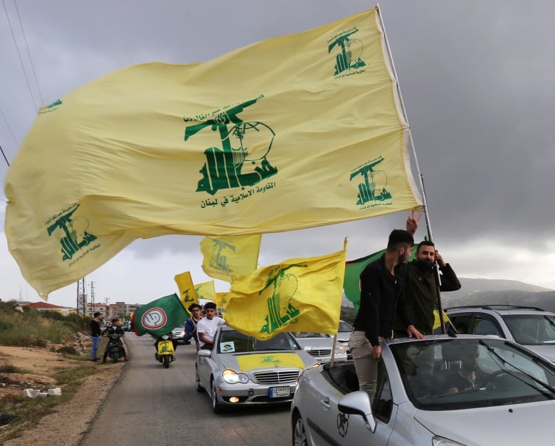 Large yellow and green flags wave from a line of cars, driving through an arid landscape. 