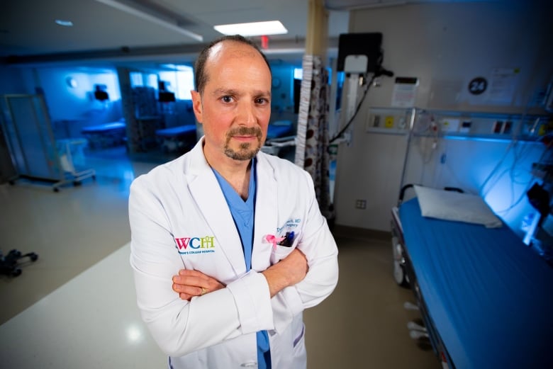 A doctor in a white coat stands in a hospital corridor looking at the camera with his arms crossed.