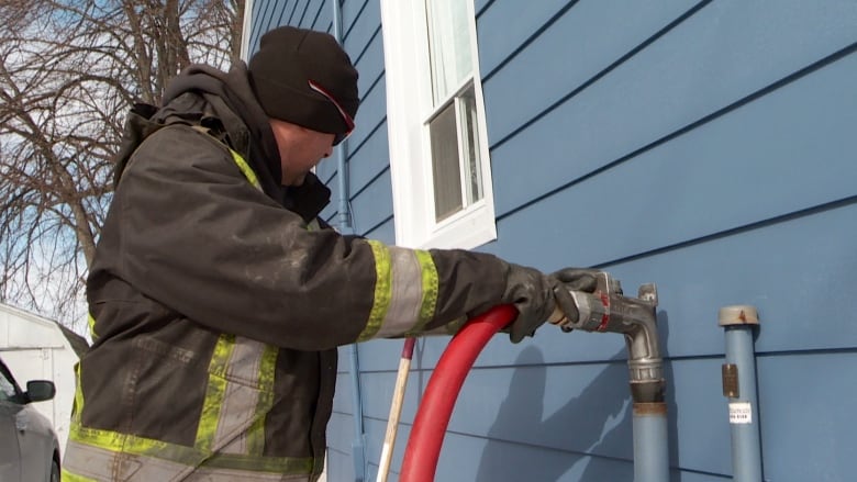 A man in a heavy coat delivers home heating oil from a large hose to an intake pipe on the side of a house in P.E.I. 