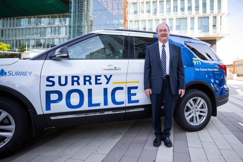 A smiling white man in a dark-blue suit stands beside an SUV emblazoned with the words 