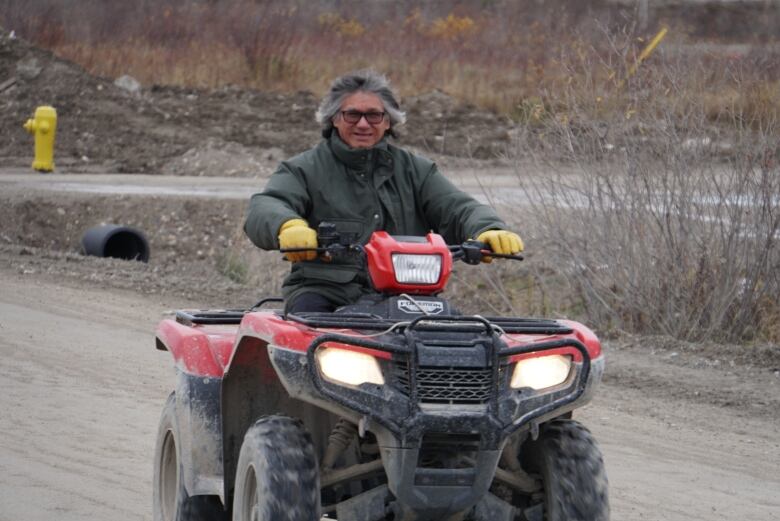 A man with grey hair rides a red ATV along a dirt road with a fire hydrant in behind him 