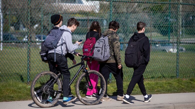 Group of students walking by a school fence. One student is sitting on a bike and looking at his phone.