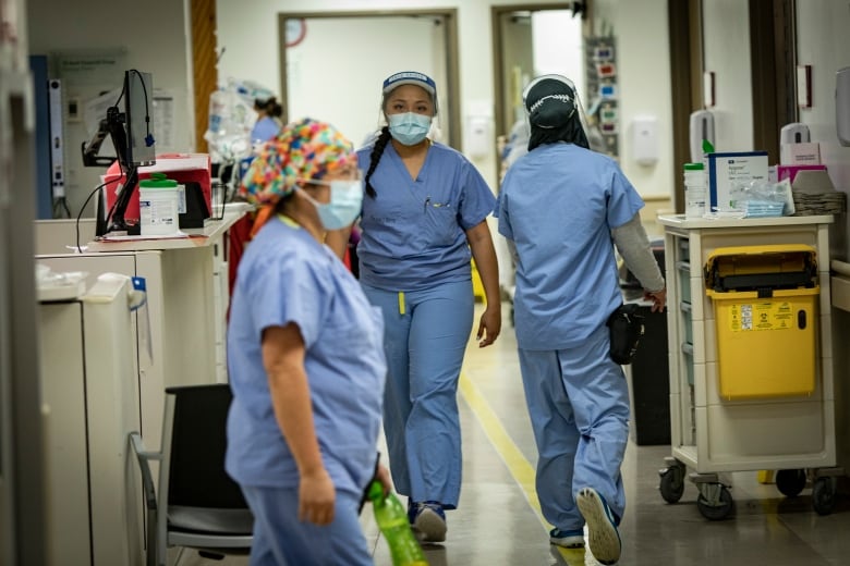 Three nurses, masked and wearing blue scrubs, walk in different directions through a hospital corridor. 