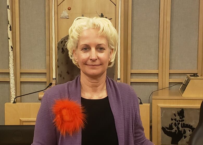 A smiling woman wearing a purple sweater stands in a territorial legislature.