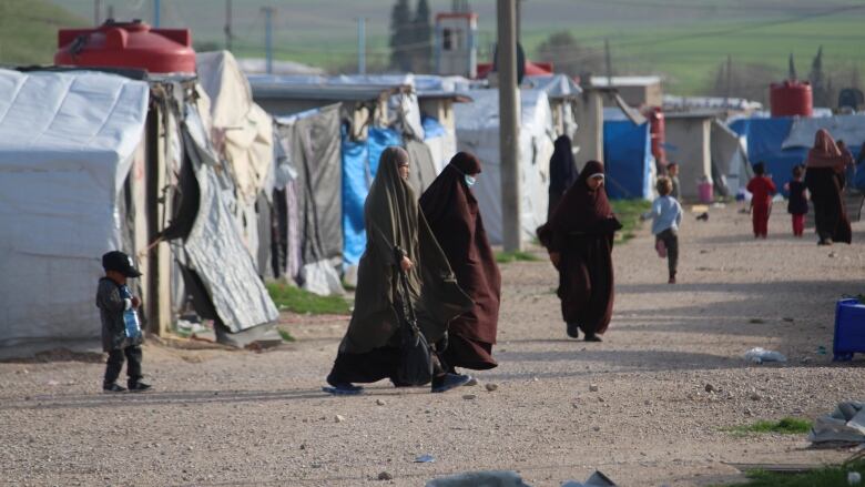 three women in head to toe Islamic coverings walk among children on a gravel road flanked by tents