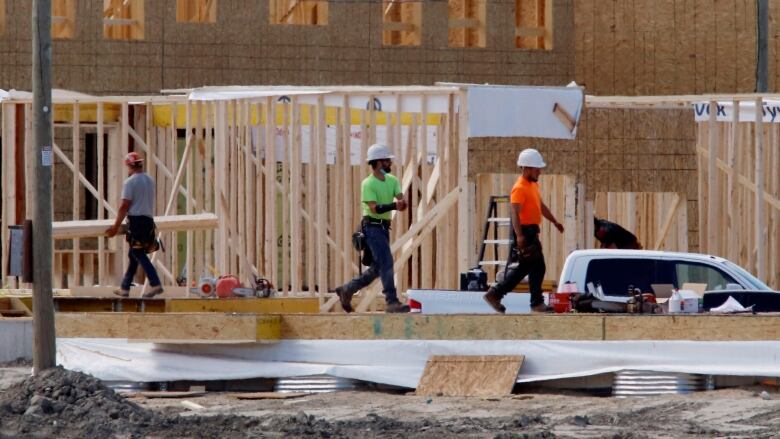 Three men dressed in hard hats and brightly coloured shirts walk in front of a partly constructed home.