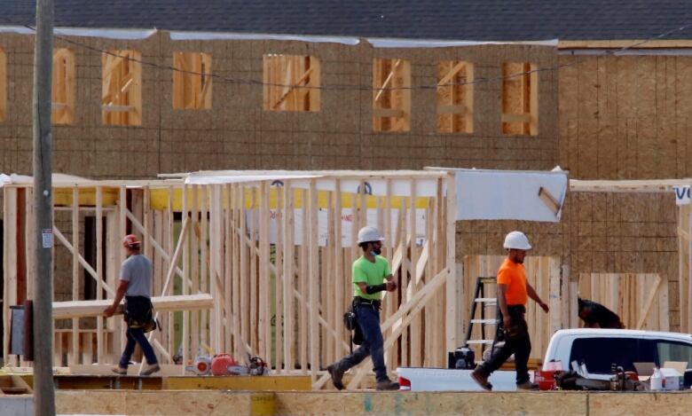 Three men dressed in hard hats and brightly coloured shirts walk in front of a partly constructed home.