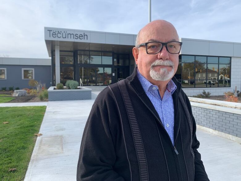 A man with a goatee and reading glasses poses in front of a municipal building. 