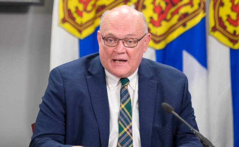 A man is shown speaking in front of a Nova Scotia flag.