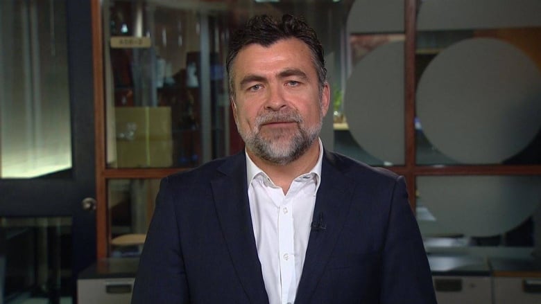 A man with a blue suit and thin grey beard looks into the camera.