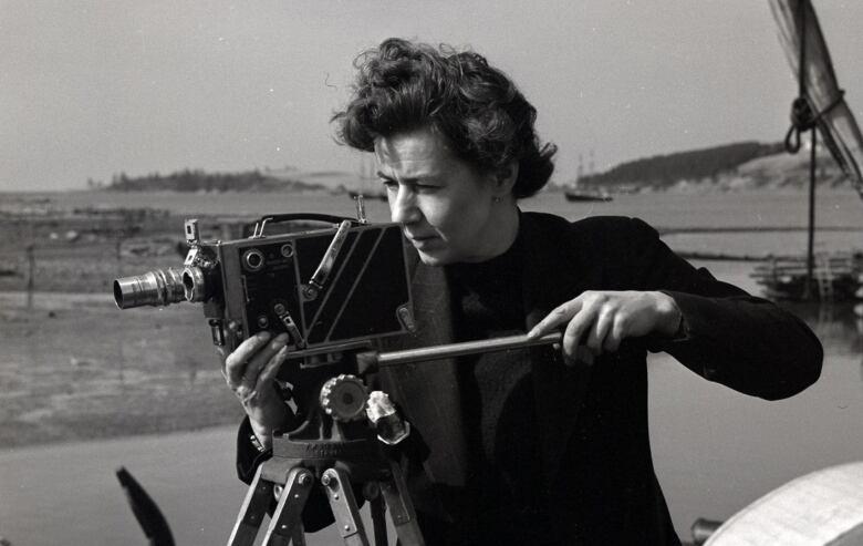 A black-and-white photo of a woman operating a film camera atop a tripod. 