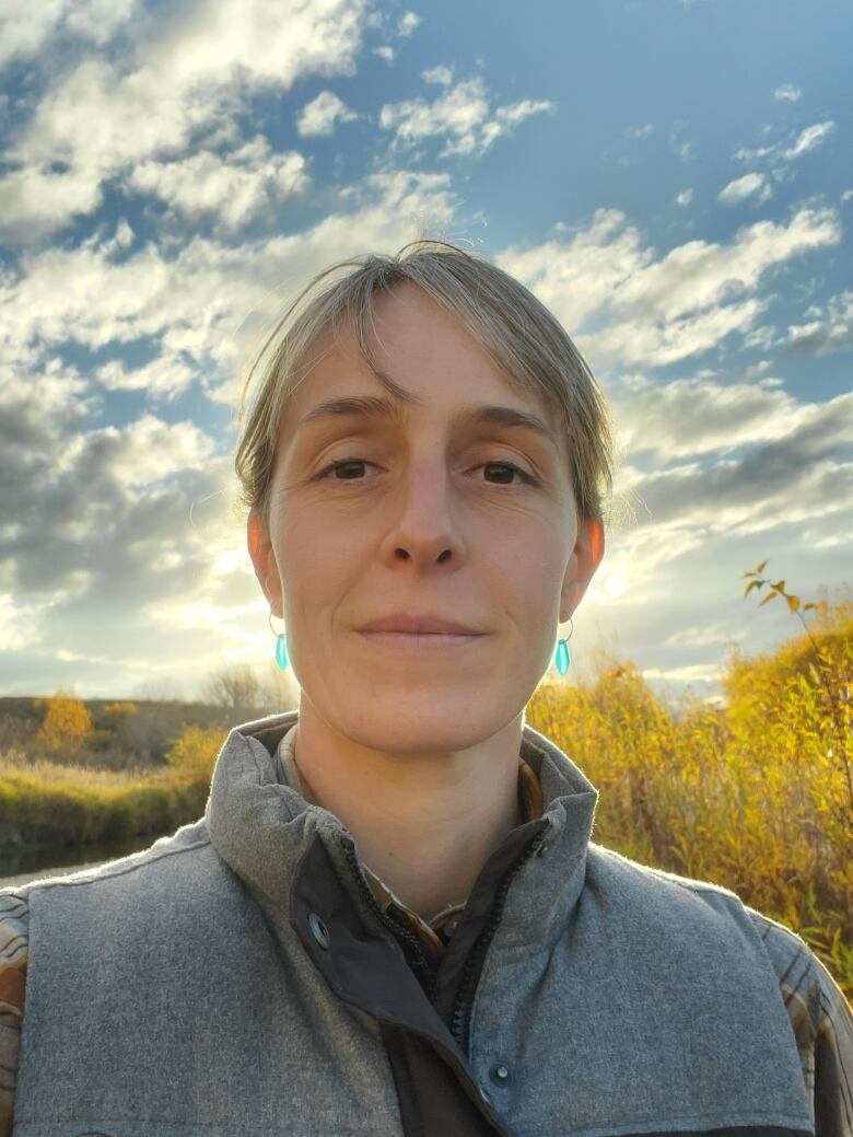 A woman takes a selfie on a fall day. There are trees behind here that are yellow. She has blonde hair in a ponytail and is wearing blue earrings. 