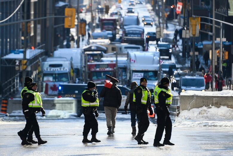 A small group of police officers in yellow vests walk past a long line of trucks parked on a city street. Someone carries a fuel can past the officers.