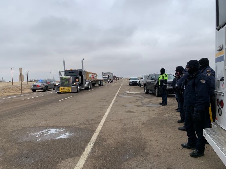 A line of semi trucks stretches down one side of a highway while R-C-M-P observe.