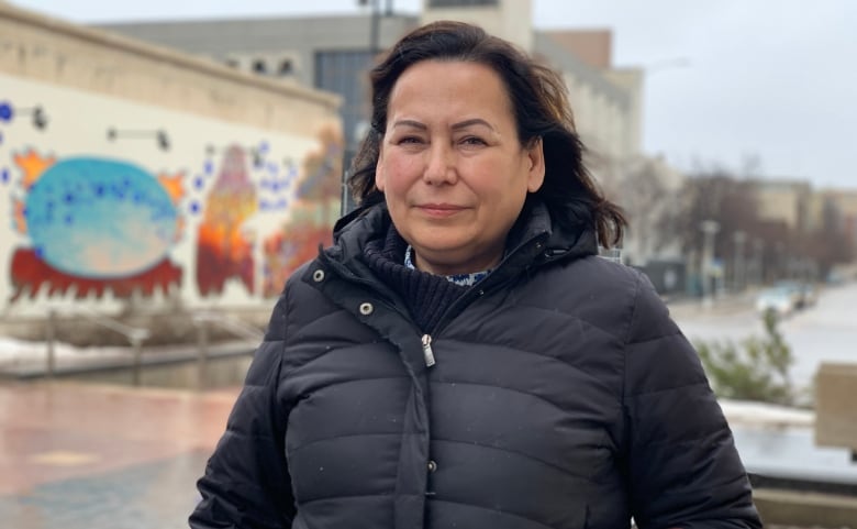 A woman with long black hair and wearing a black puffy winter jacket stands outside.