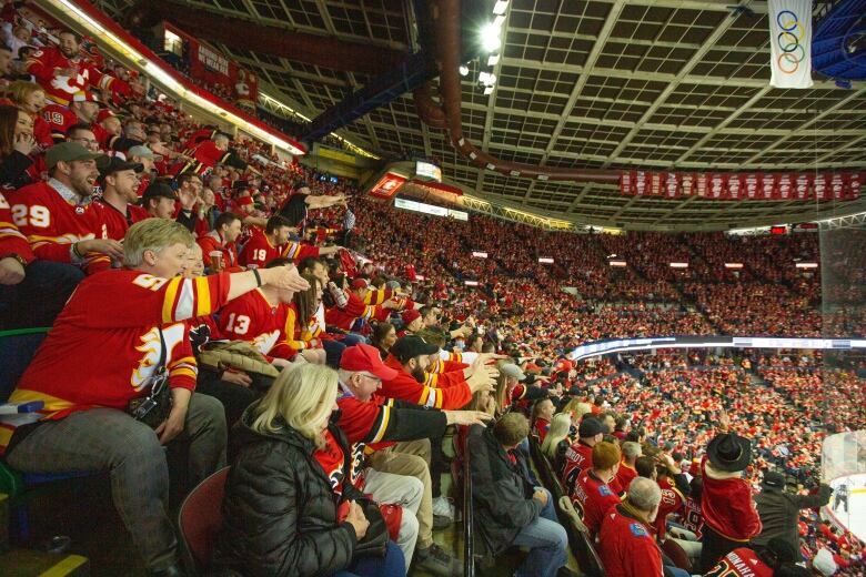 A large crowd of fans mostly dressed in red Calgary Flames jerseys are cheering in their seats at the Calgary Saddledome arena.