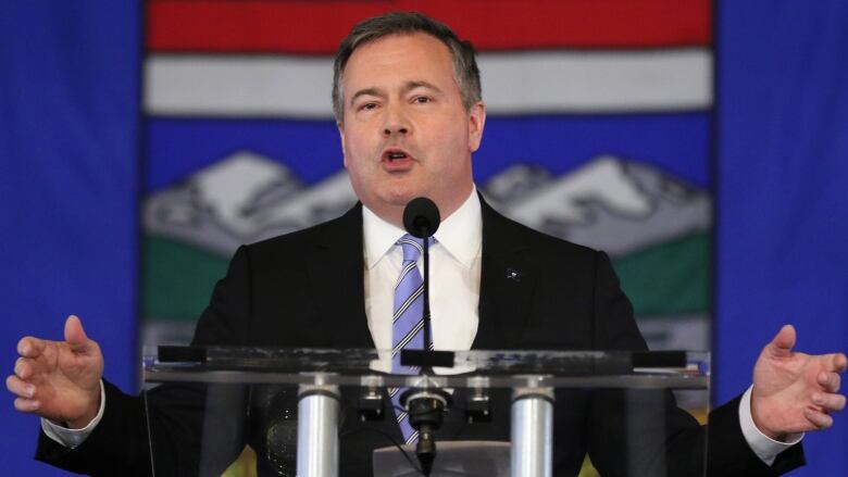 A man wearing a suit gives a speech with the Alberta flag in the background.