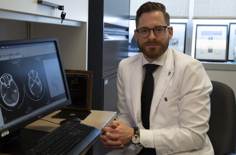 A doctor in a white coat sits next to a computer monitor showing a brain scan.