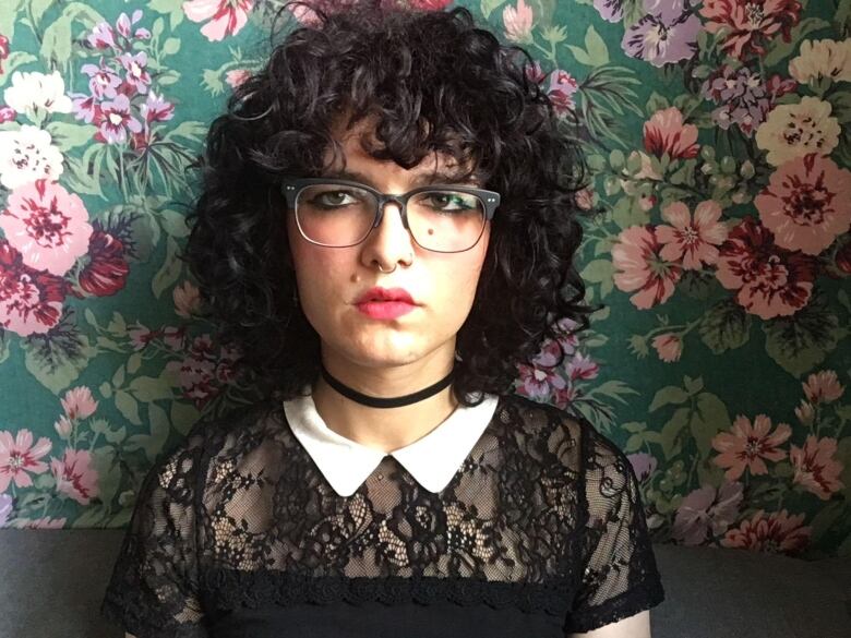 A woman with curly black hair and glasses posing in front of a flowery wallpaper.