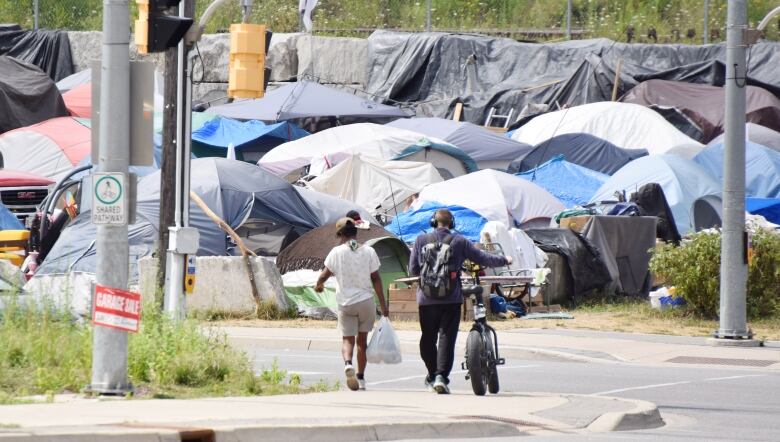 Two people, one walking beside a bicycle, cross at an intersection and walk towards multiple tents.