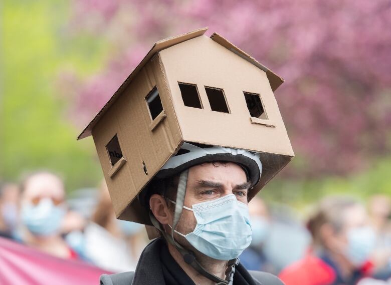 A man with a face mask wears a cardboard house on his head atop a bike helmet. 