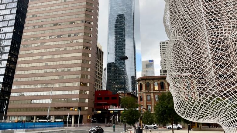 Tall buildings and downtown street. In the foreground is a white wire sculpture of a man's face.