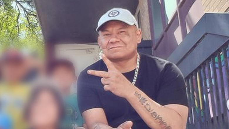 An Indigenous man with a Winnipeg Jets hat on makes the peace symbol towards the camera. He has tattoos all along his arms.