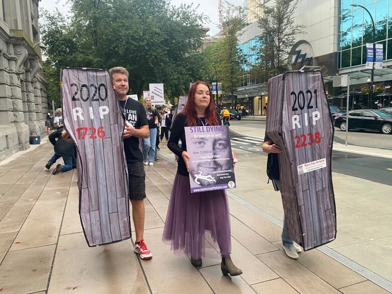 Two people holding coffins with the toxic drug death toll listed on them walk next to a woman with a sign saying 'Still Dying' and a picture of a dead loved one.