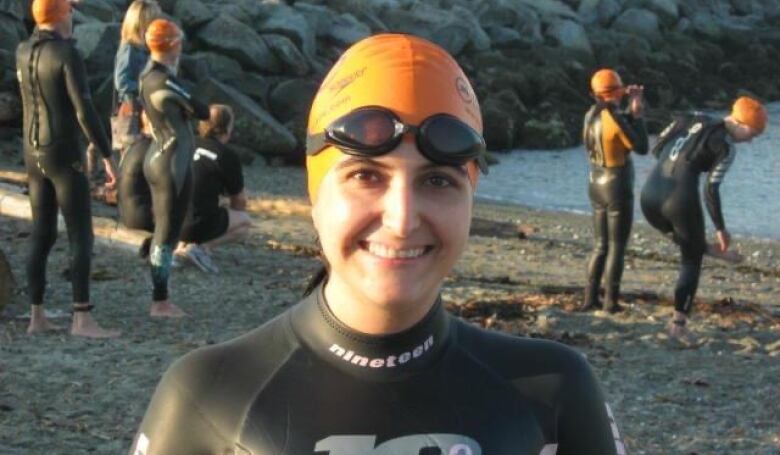 A woman in diving gear, with an orange swimming cap on, stands on a beach surrounded by similarly-dressed people.