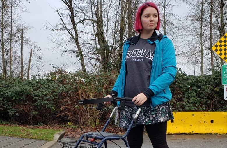 A woman with pink hair stands with a walker on a street.