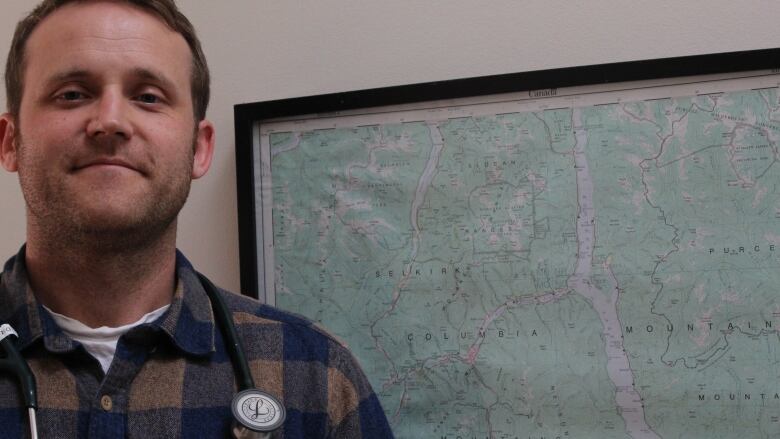 A doctor smiles in front of a map. He is wearing a checked shirt and has a stethoscope on.