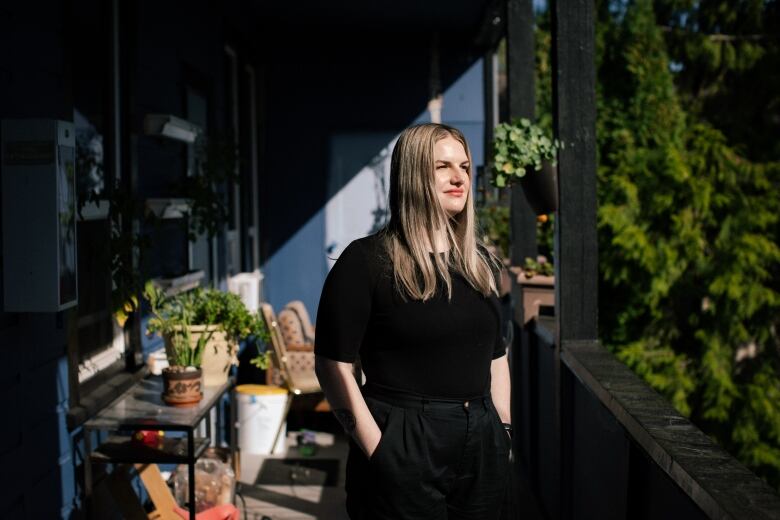 A woman dressed all in black with long blonde hair stands on a sunny balcony with potted plants behind her. She has her hands in her pockets and is looking out of frame, to the right.