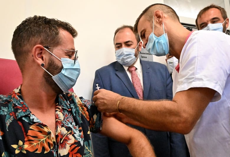 Person wearing tropical short-sleeved shirt and medical face mask sits as a health professional wearing a mask holds a vaccine needle above his arm, while others watch.