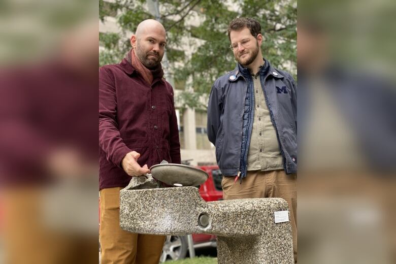 Two men stand behind a broken water fountain.