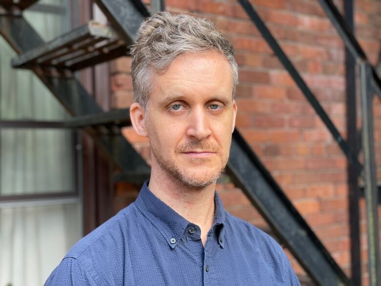 A slender grey-haired white man in a blue button-up collared shirt looks directly at the camera with a neutral facial expression.