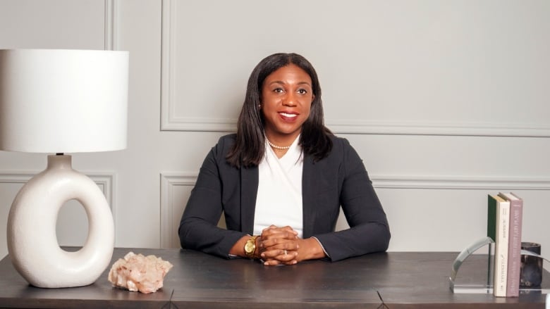 A Black woman with shoulder-length hair sits at a large wooden desk looking straight ahead, wearing a white top, black blazer, and her hands are folded on the desktop.