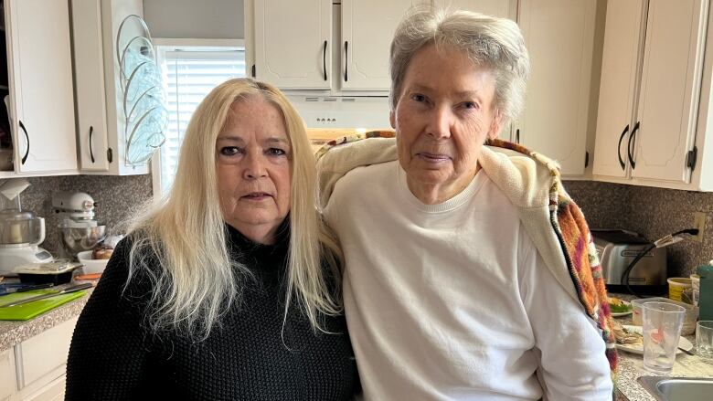 Two older white women stand in a kitchen facing the camera with their arms on each other's shoulders. The woman on the left has long straight blonde hair and is wearing a black sweater, while the woman on the right has short silver hair and a long-sleeved white t-shirt.