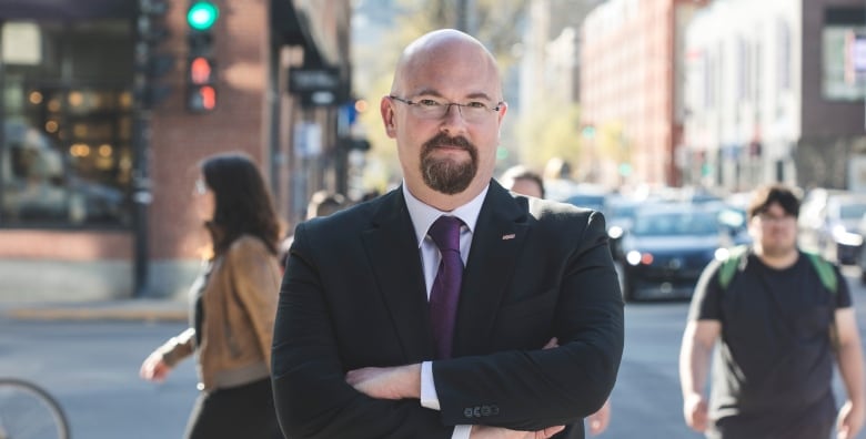 Bald man in suit standing on a busy sidewalk with arms folded