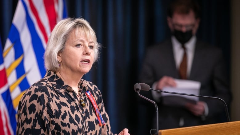 Dr. Bonnie Henry, a white woman with short white hair, talks at a podium while Adrian Dix, wearing a mask, stands in the background.