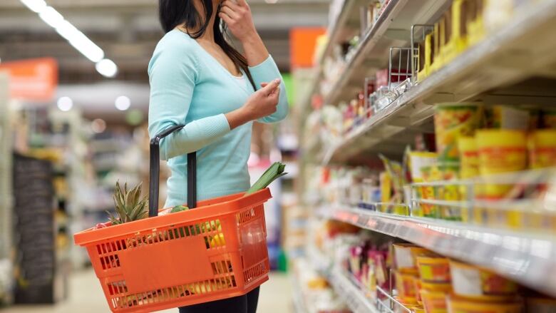 A woman with a small basket in her hand looks at items on the shelves of a grocery store.