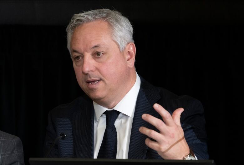 A man in a dark suit speaks into a microphone.