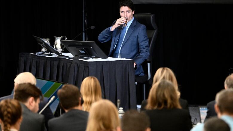 A politician drinks from a water glass as he looks out at people seated in a hearing room.