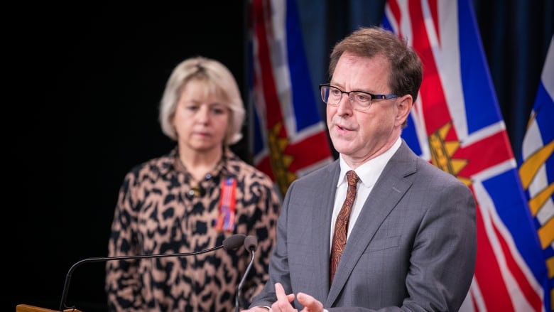 A man speaks at a podium, with the flag of British Columbia behind him. A woman with short hair stands in the background.